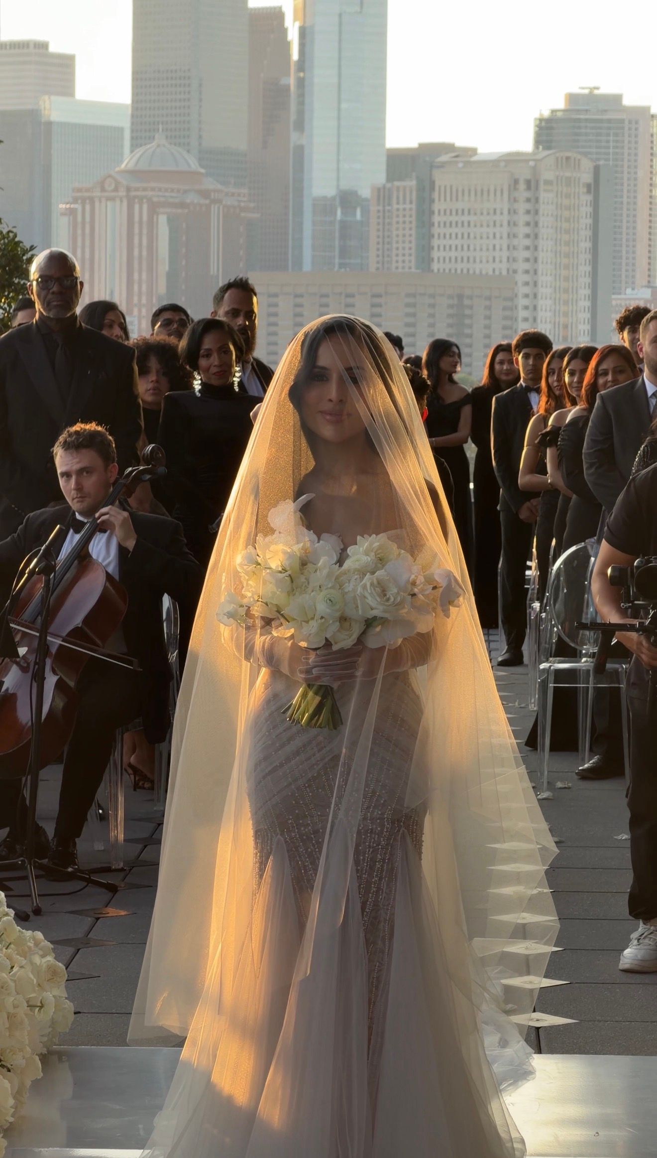Bride in a fitted lace gown with a long veil holds white roses bouquet, walking down the aisle at an outdoor rooftop wedding.