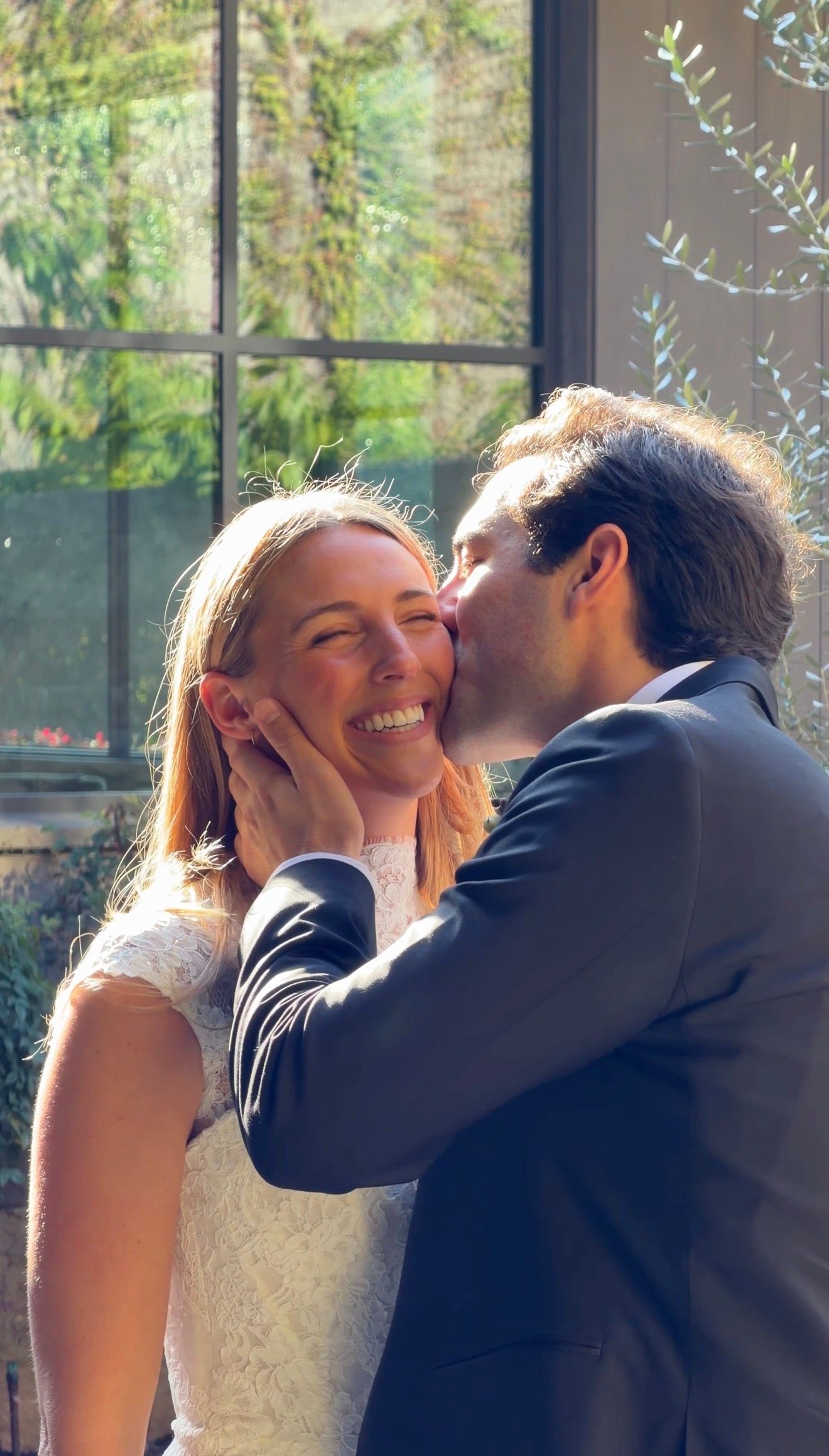 Groom in blue suit kisses smiling bride in white lace dress on the cheek during wedding celebration.