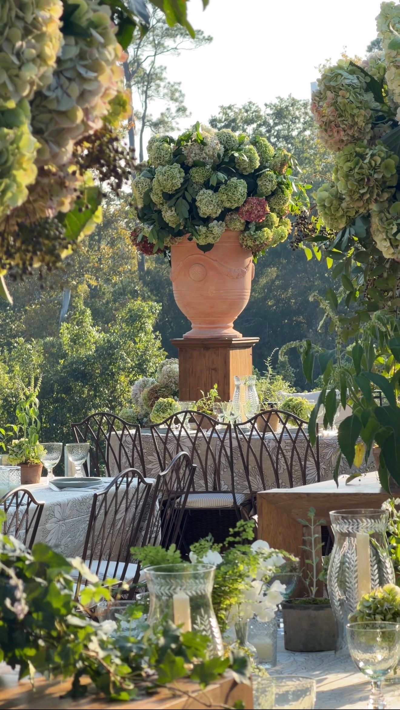 Elegant outdoor dining table with crystal glasses surrounded by lush greenery and a large terracotta urn with white hydrangeas.
