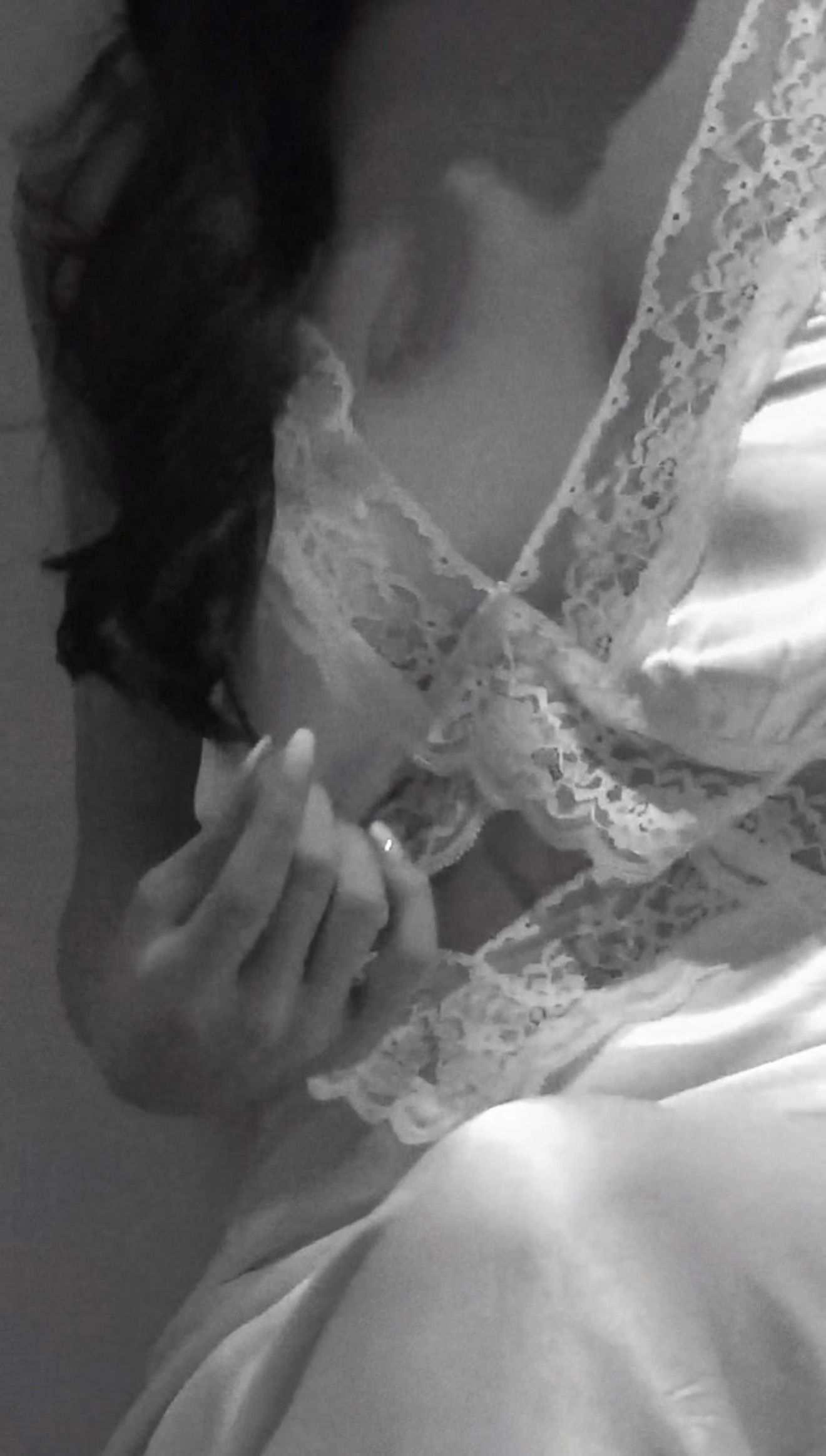 Black and white photo of a woman in elegant white lace lingerie, holding a small object with manicured nails.