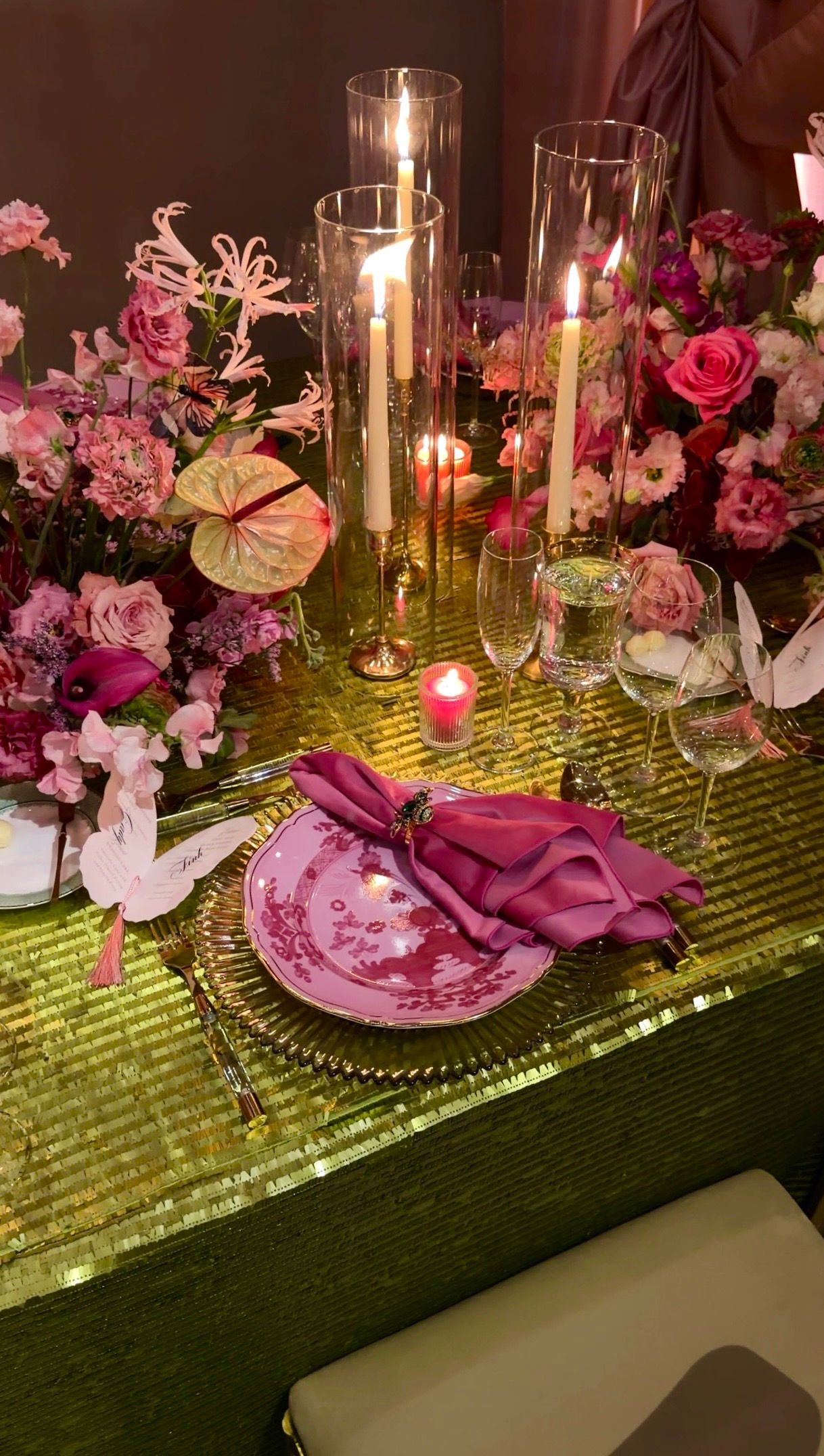 Elegant wedding table setting with pink floral arrangements, lit candles, crystal glasses, and a magenta napkin on sequin green tablecloth.