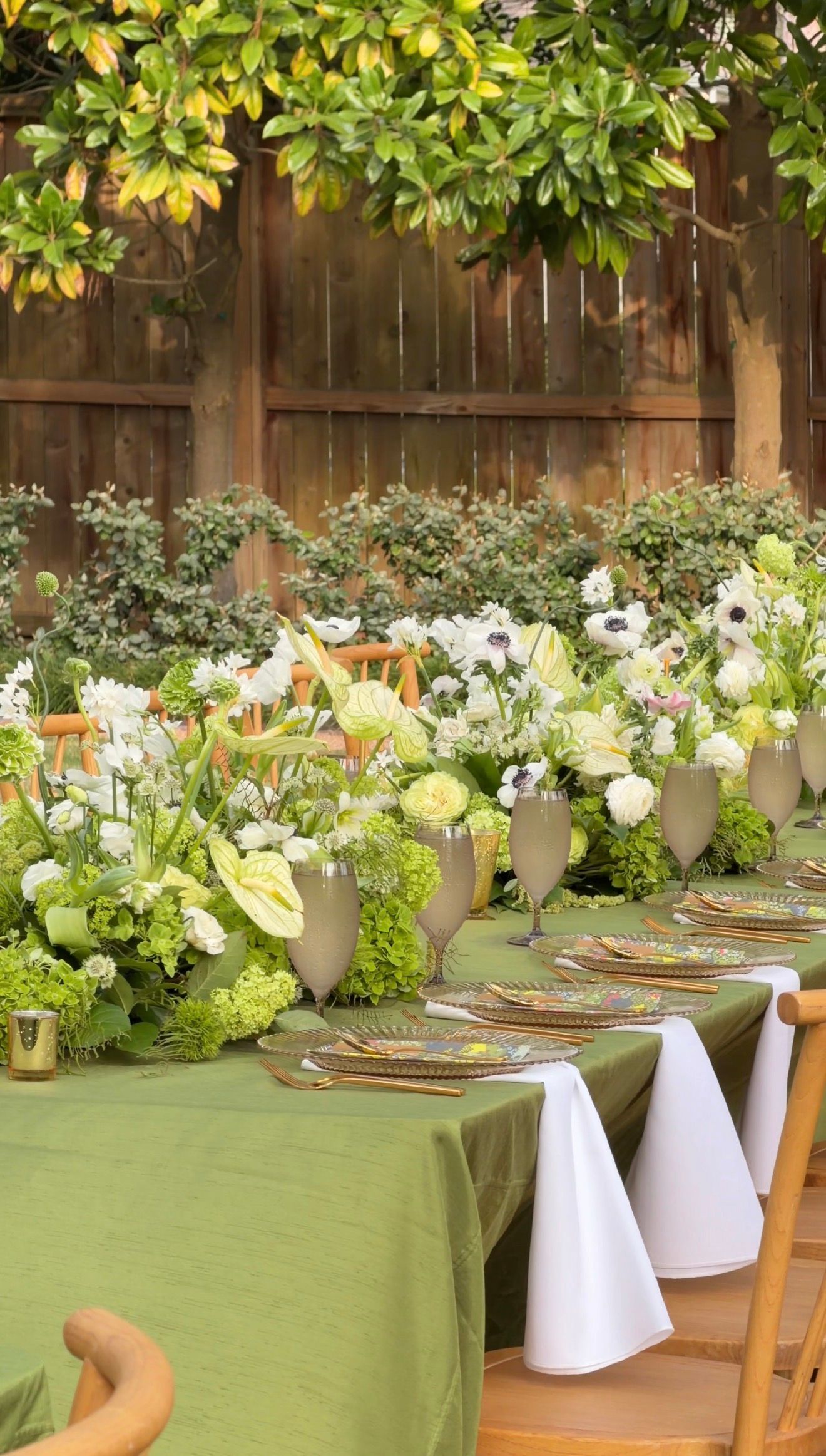Elegant outdoor dining table with green linen, floral centerpieces of white blooms, and gold chargers set in a garden.
