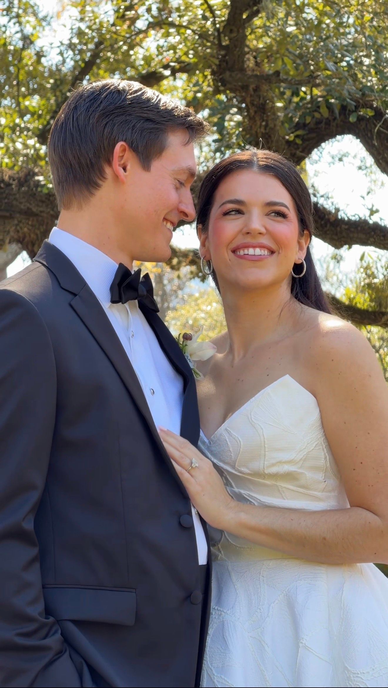 A smiling bride in a strapless white gown and groom in a black tuxedo share a romantic moment outdoors under a tree.