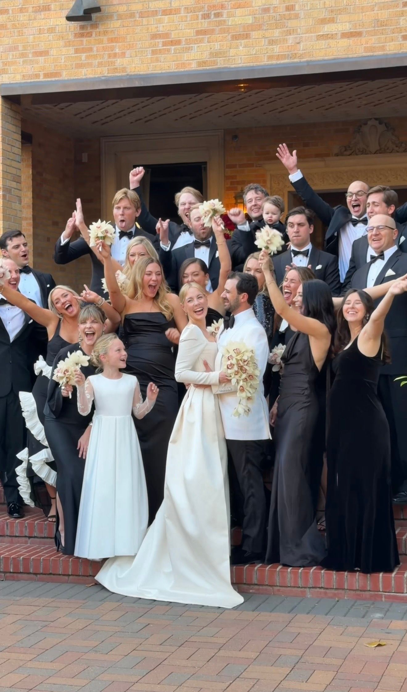 Joyful wedding party in black and white attire celebrating on church steps with bouquets raised.