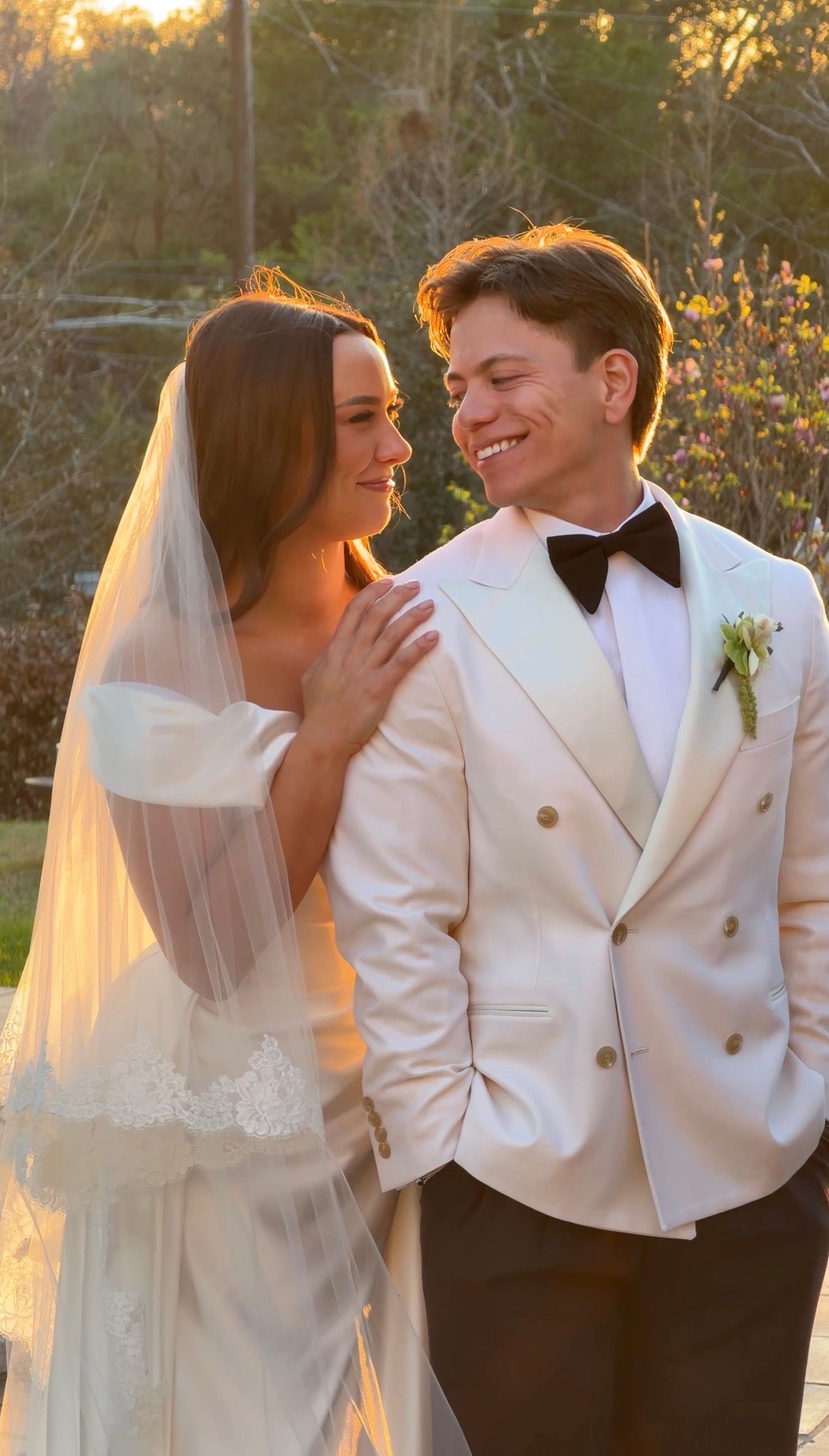 A bride in a white gown and veil smiles with a groom in a cream tuxedo with bow tie at a golden hour outdoor wedding.
