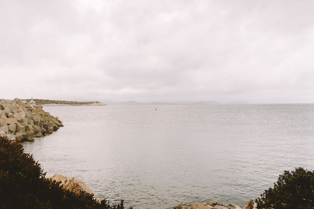 Rocky coastline meets calm gray ocean waters under an overcast sky, with a stone jetty extending into the sea.