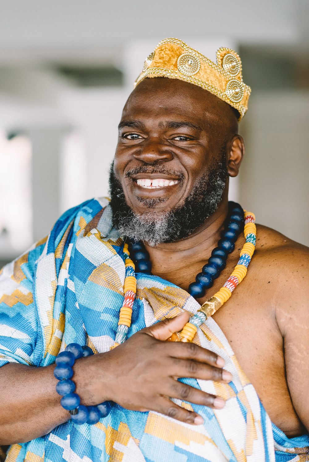 Smiling man in traditional African kente cloth, gold crown, and black beaded necklace posing regally.