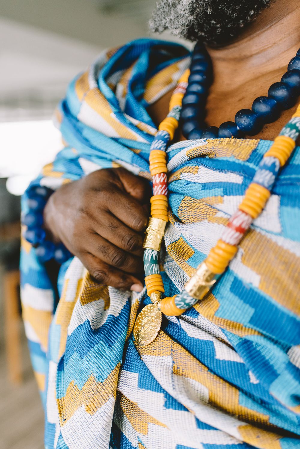 Person wearing colorful Kente cloth with blue and yellow beaded necklaces, showcasing traditional African attire.