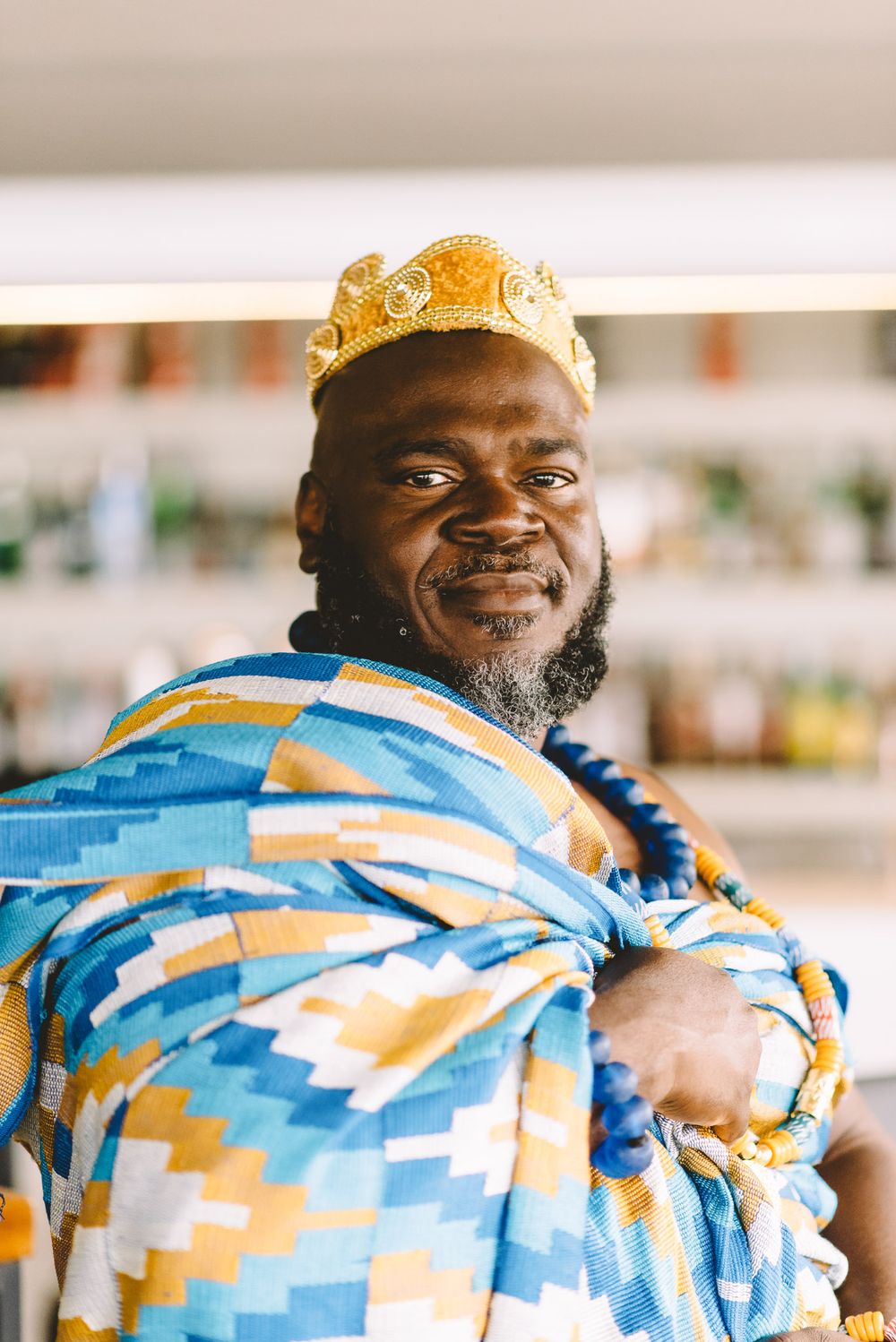 Man wearing golden crown and colorful kente cloth traditional African attire smiling confidently indoors.