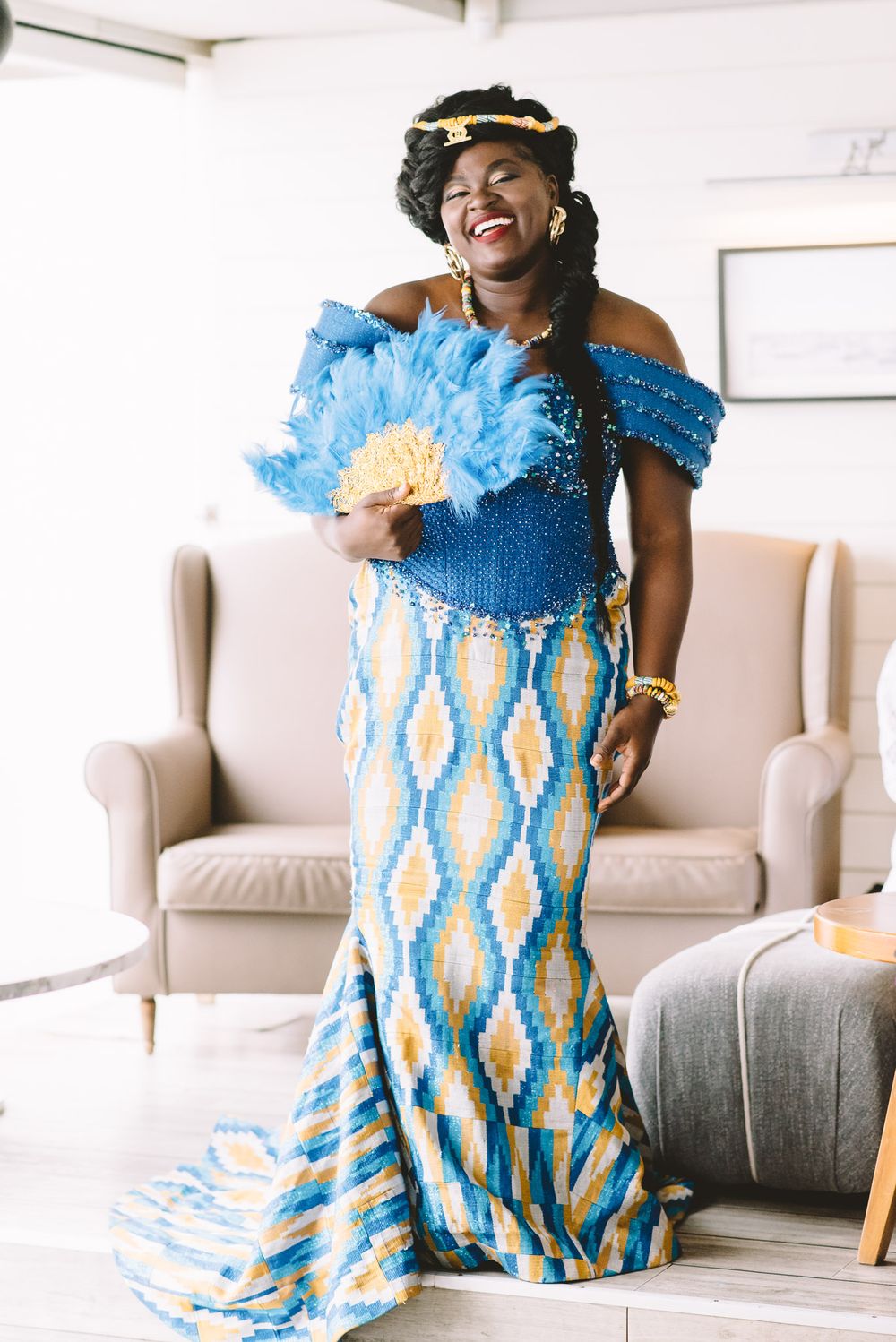 Woman in blue kente dress with feather fan smiling joyfully at traditional African ceremony.