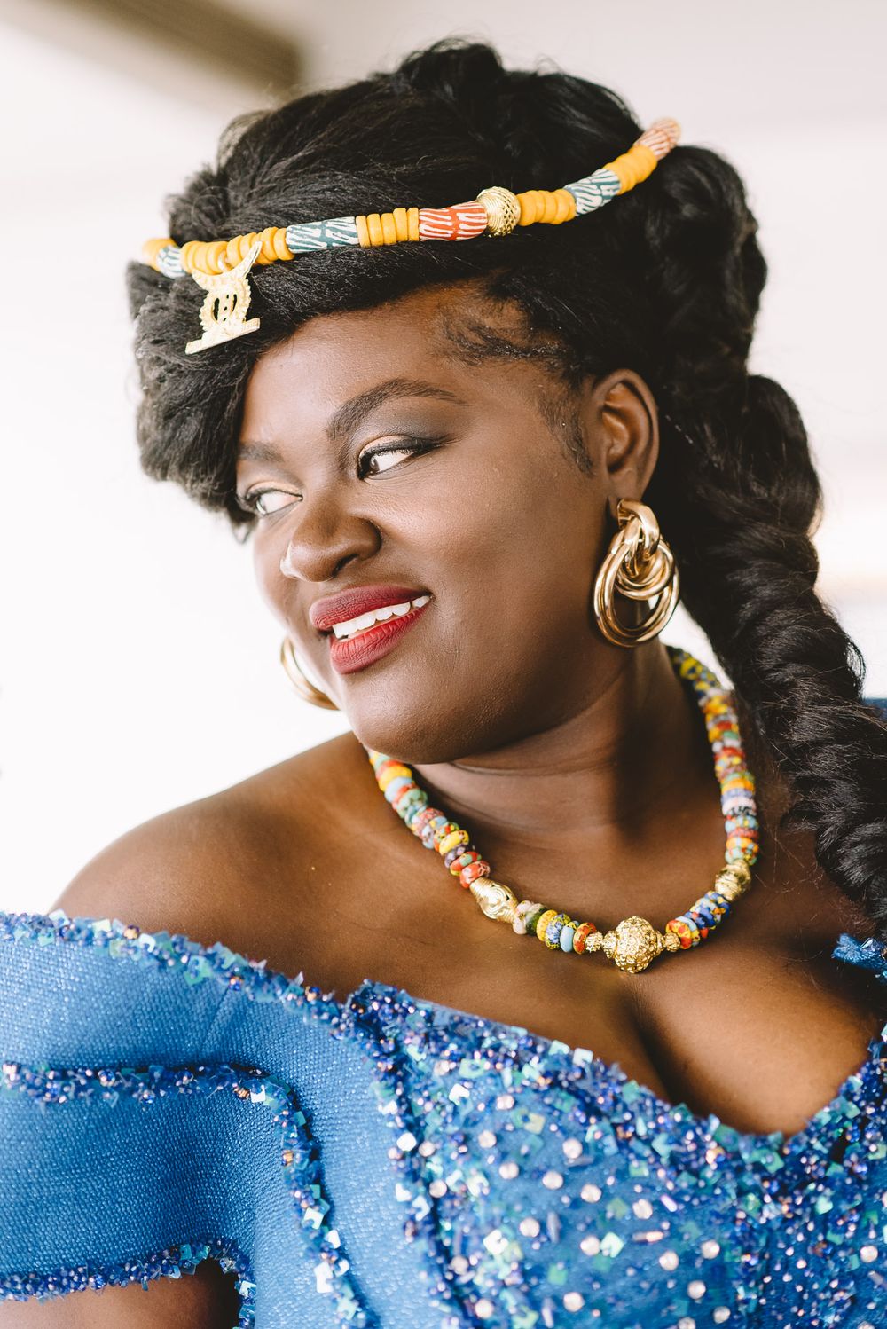 Woman in blue beaded gown with colorful hair accessory, hoop earrings, and beaded necklace smiling elegantly.