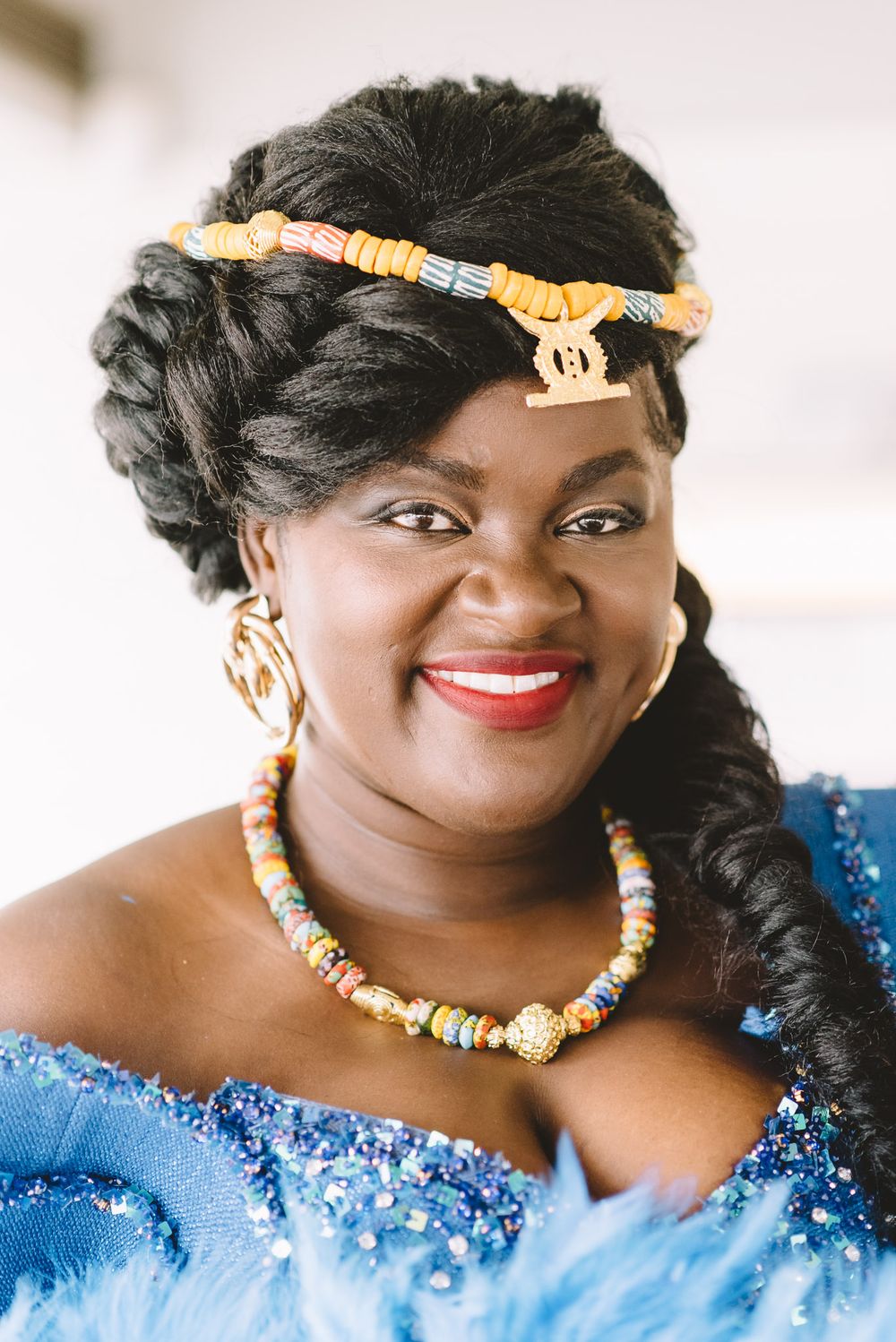 Smiling woman in blue sequin dress with colorful beaded headpiece, necklace, and gold earrings in traditional African attire.
