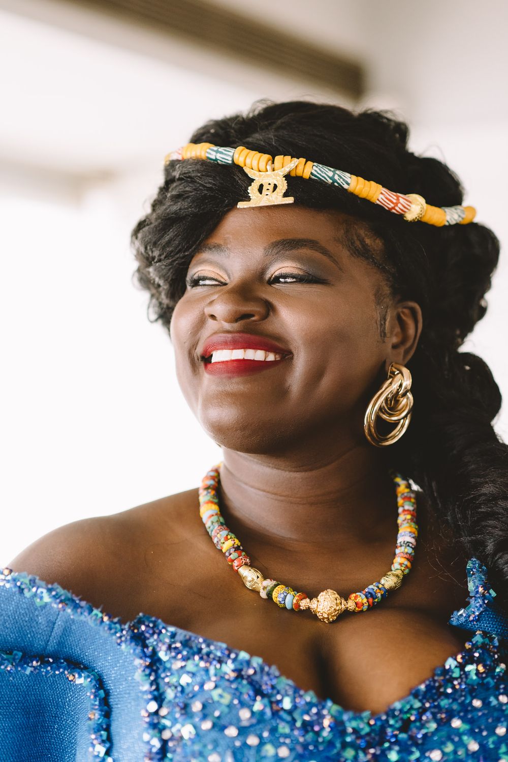 Smiling Black woman wearing blue sequin dress, colorful beaded necklace, gold hoop earrings, and yellow headband.
