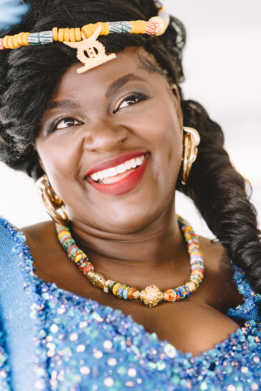 Smiling woman wearing colorful beaded jewelry, gold earrings, and a sparkling blue dress with traditional African accessories.