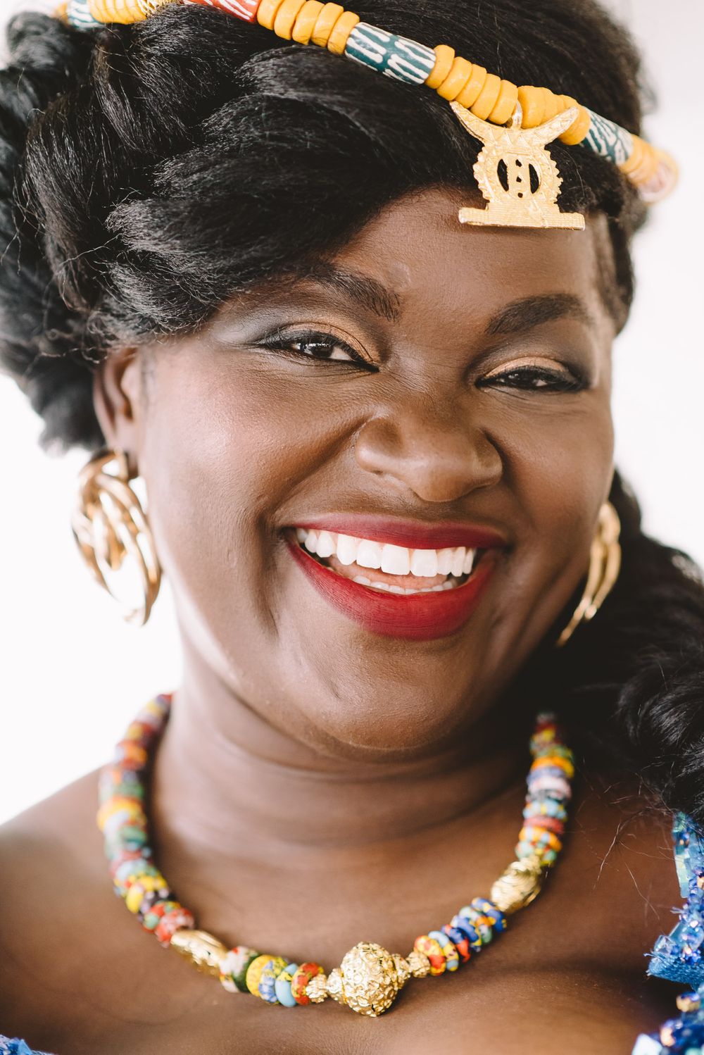 Smiling African woman wearing colorful beaded necklace, gold earrings, and traditional hair accessories with red lipstick.