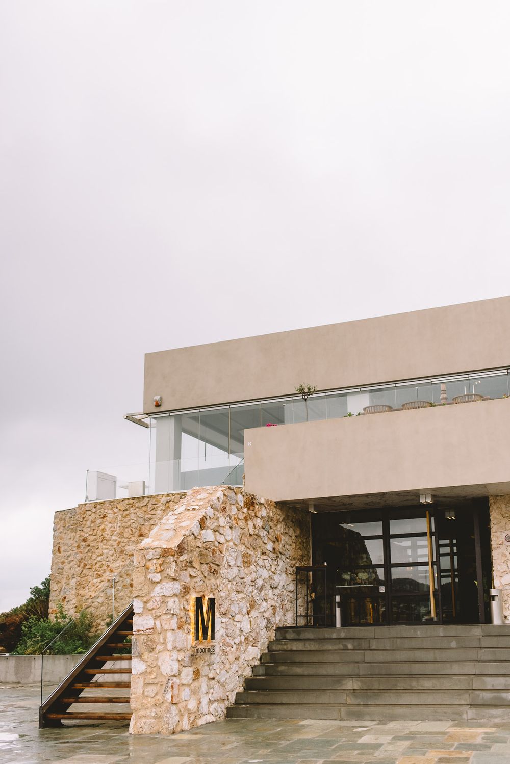 Modern stone and concrete building entrance with glass railings and letter M sign on a overcast day.