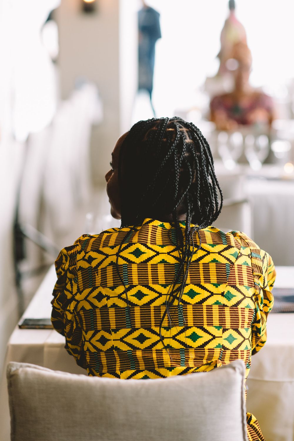 Person seen from behind wearing vibrant yellow and green African kente cloth shirt with braided hair.