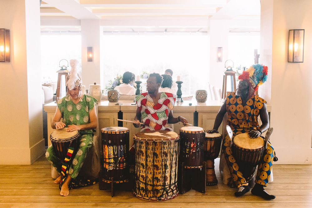 Three musicians in colorful traditional attire playing djembe drums indoors at a cultural event.