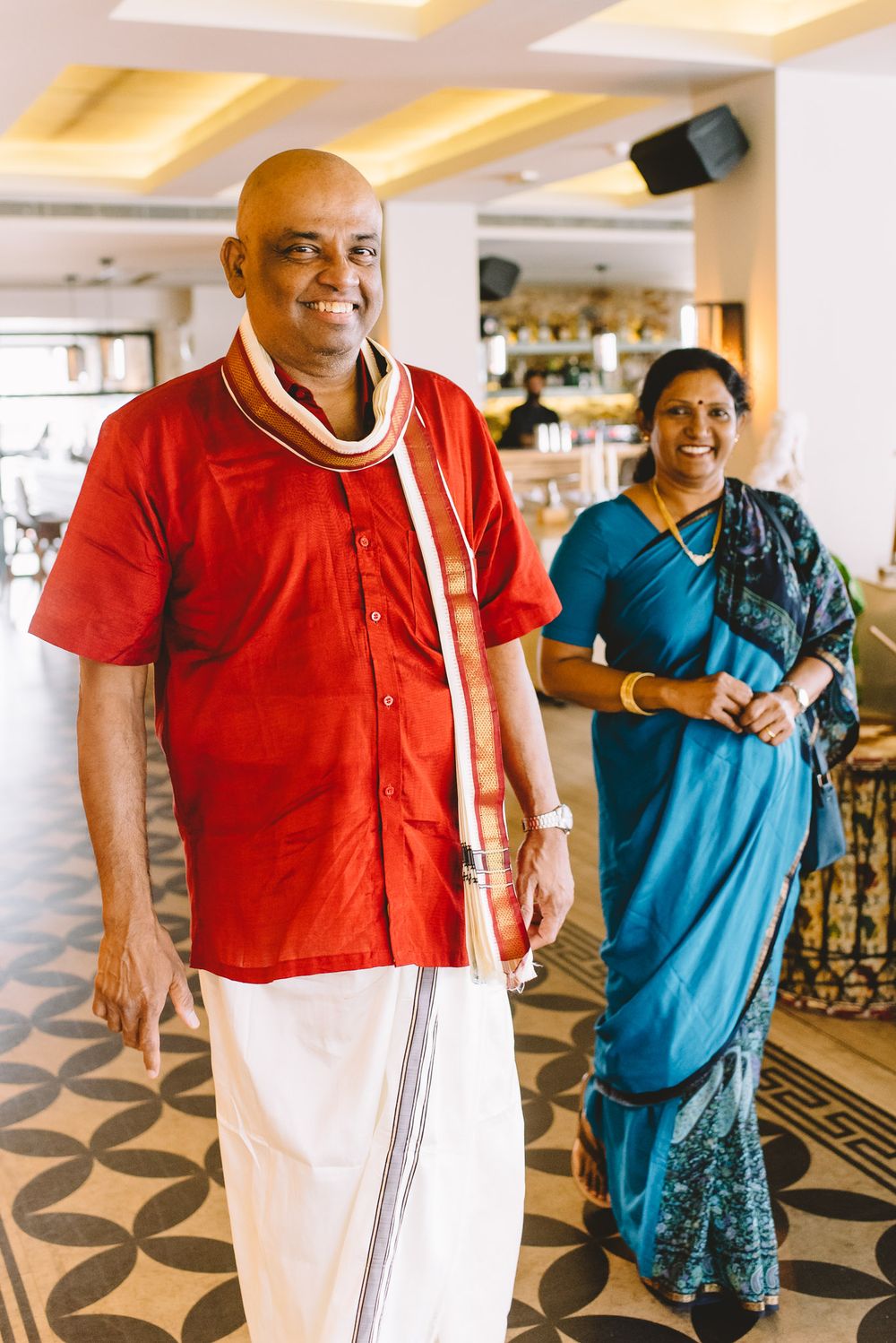 Indian couple in traditional attire, man in red kurta and white dhoti, woman in teal saree, smiling indoors.