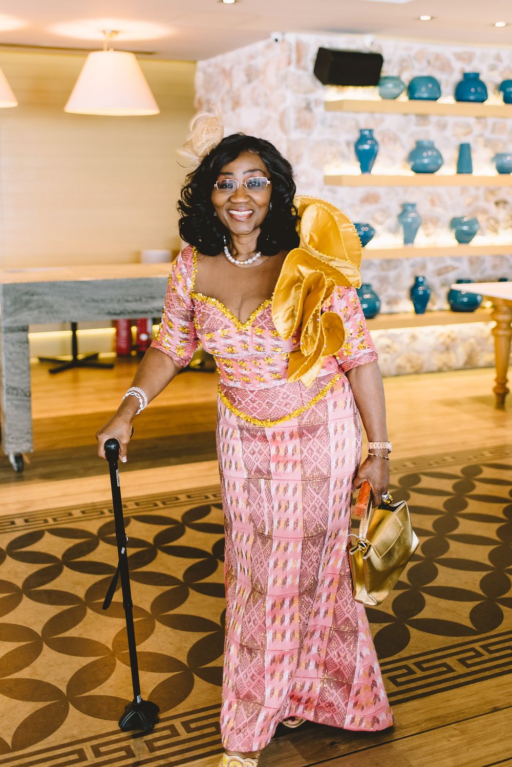 Woman in pink traditional African dress with gold accessories and cane poses indoors.