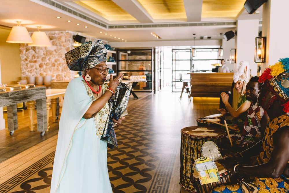 Woman in traditional African attire and headwrap speaks at a cultural ceremony in a warmly lit restaurant.