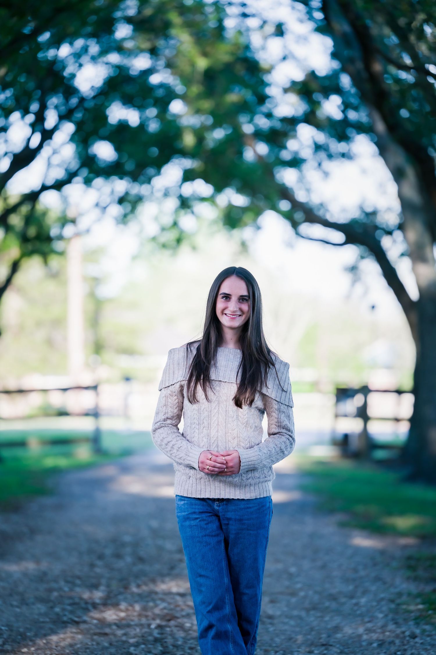 Young woman in beige cable-knit sweater and jeans smiling on a tree-lined gravel path outdoors.