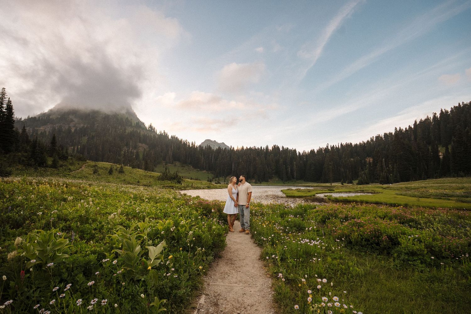 Couple embracing on a mountain trail surrounded by wildflowers, trees, and a serene lake under a dramatic cloudy sky.