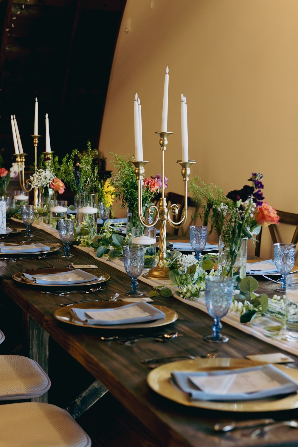 Elegant wedding reception table with gold candelabras, colorful florals, blue glassware, and gold charger plates.