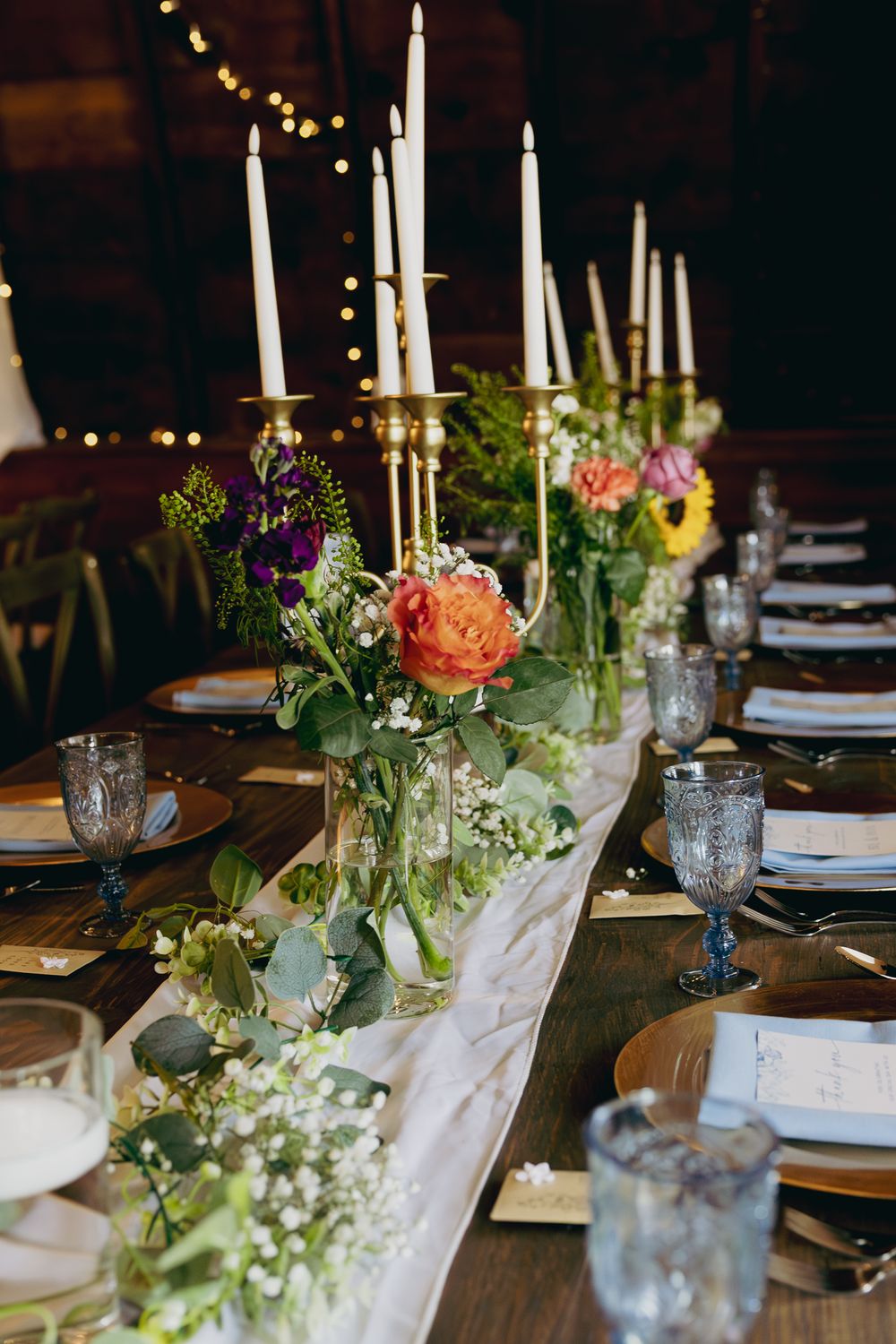 Rustic wedding table centerpiece featuring tall white candles in gold holders surrounded by vibrant fresh flowers.