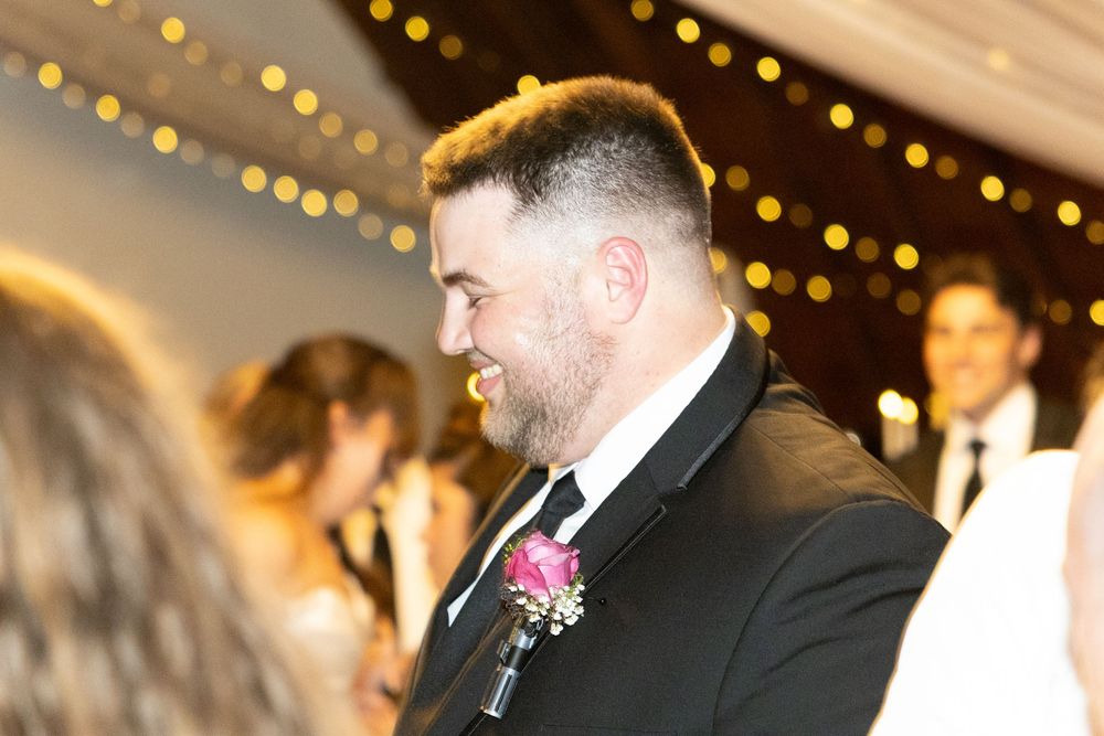 Groom in black tuxedo with pink boutonniere smiling at wedding reception with string lights.