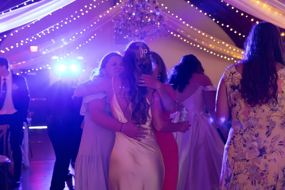 Bridesmaids in elegant gowns dancing together under purple lights at a wedding reception with string lights.