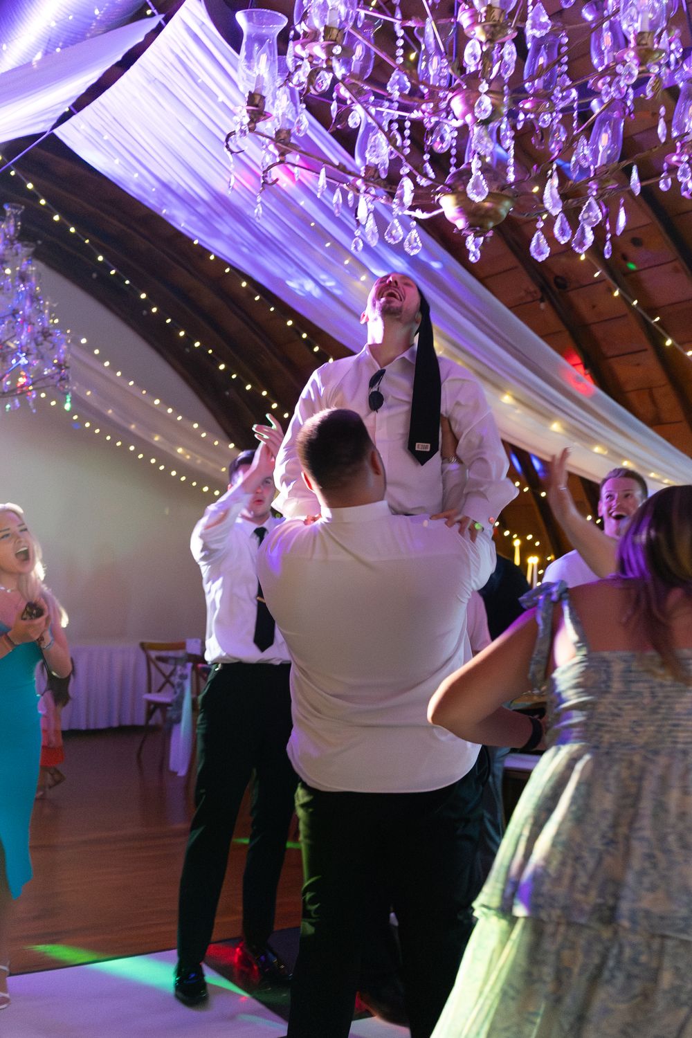 Groom lifted by guests on wedding reception dance floor under crystal chandelier with festive string lights.
