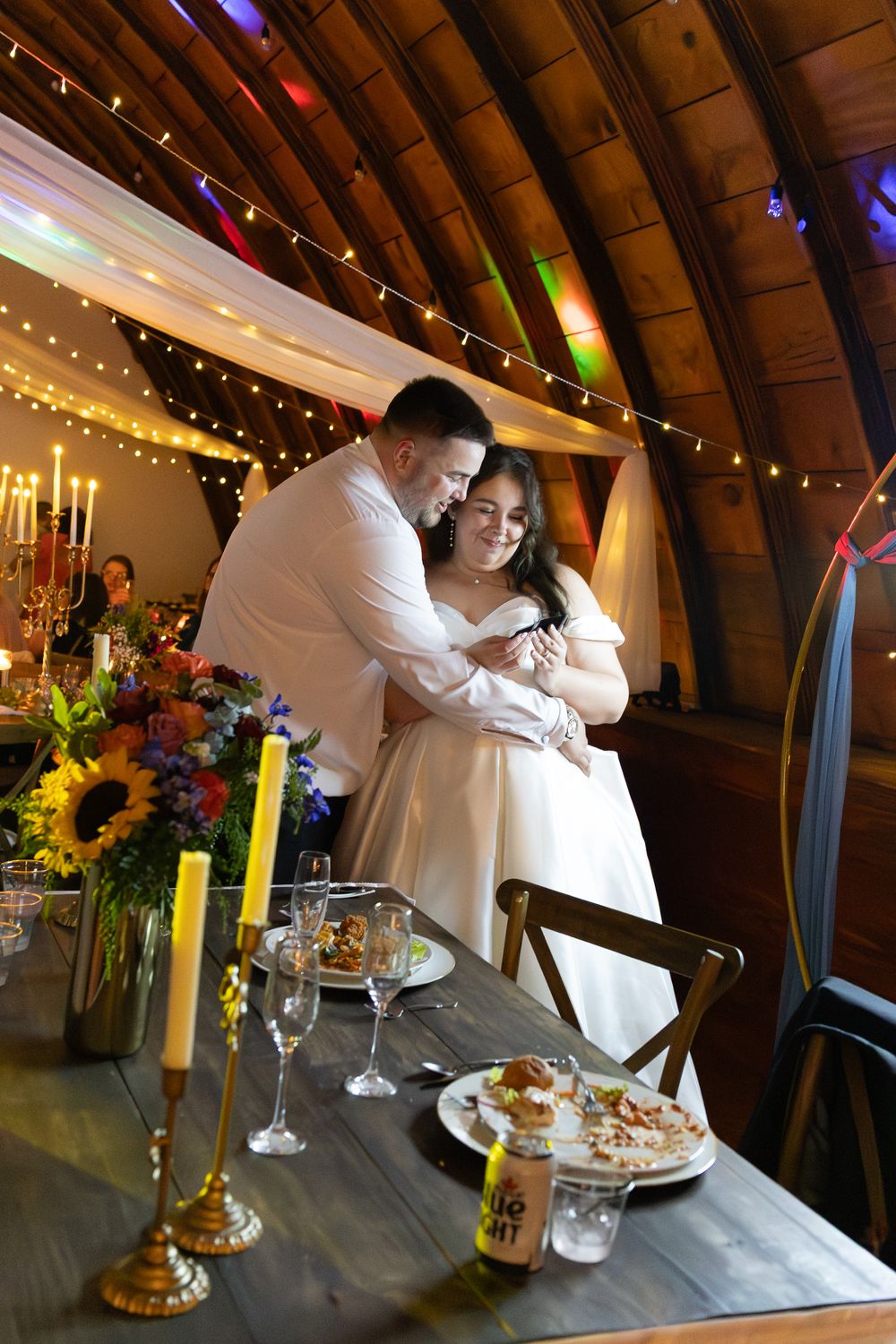 Bride and groom share a tender embrace at their sweetheart table in a warmly lit barn wedding reception.