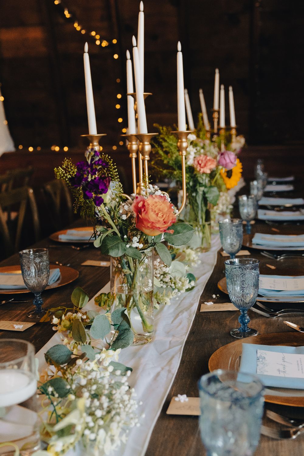 Elegant wedding table with gold candelabras, white taper candles, colorful floral arrangements, blue glassware, and gold charger plates.