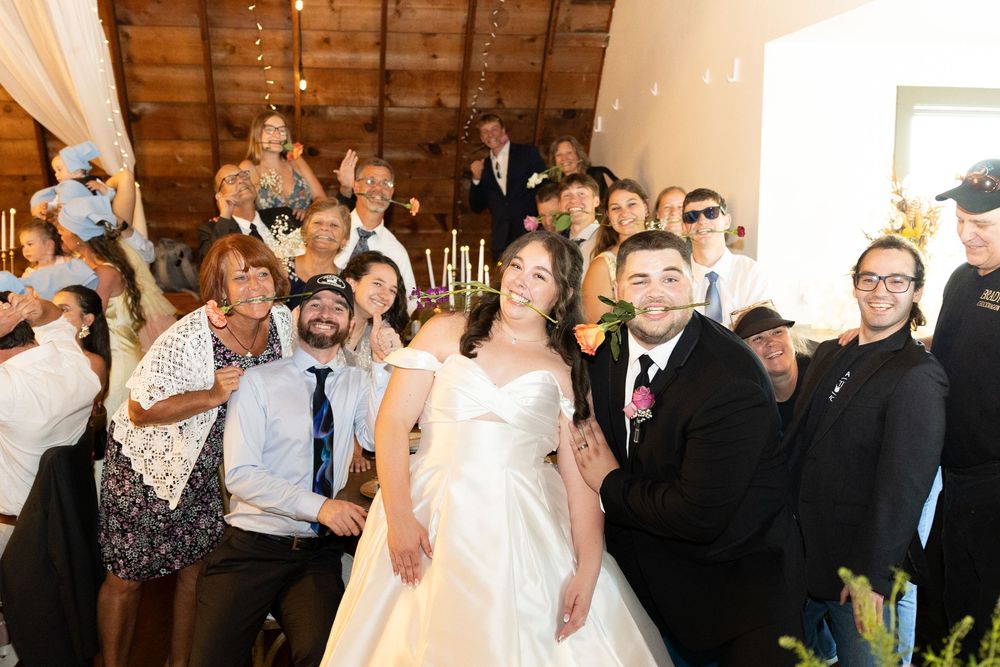 Joyful wedding couple poses with guests for a group photo inside a rustic barn venue decorated with string lights.