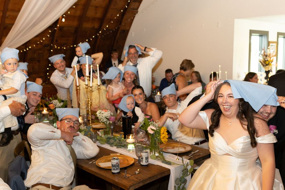 Wedding guests wearing blue paper hats celebrate at a rustic reception dinner table with candles and floral centerpieces.
