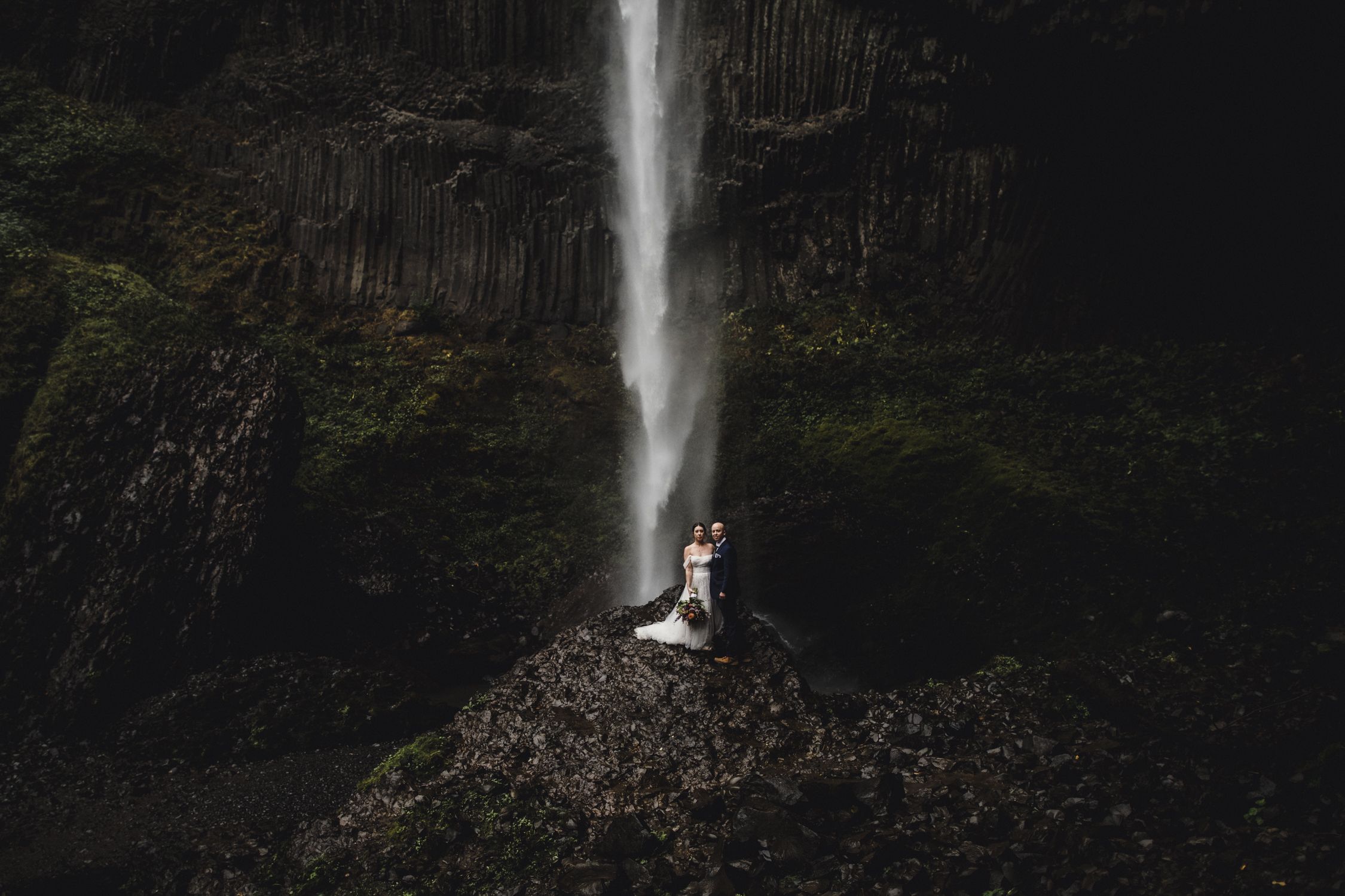 Couple embraces on rocky ledge before dramatic waterfall with dark basalt columns in moody outdoor wedding photo at Latourell Fals