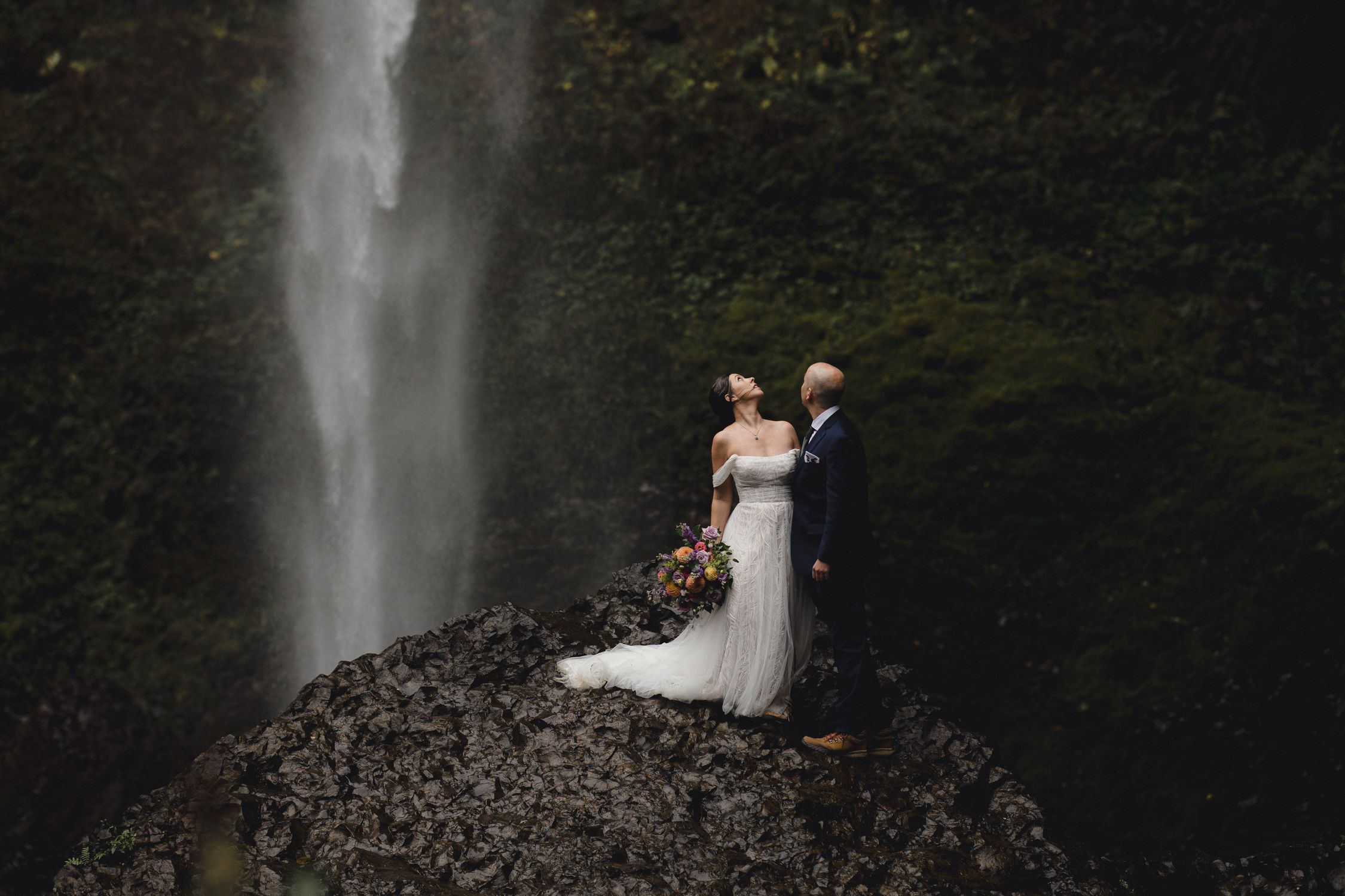Couple looks up at the 222 feet of Latourell Falls with the waterfall behind them. The background is dark from the wet basalt.
