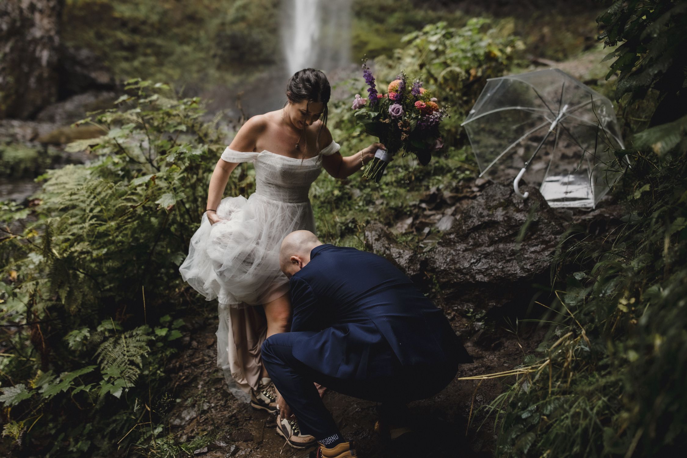 Groom ties the bride's hiking boot laces before they climb up the rocks at Latourell falls in Oregon.