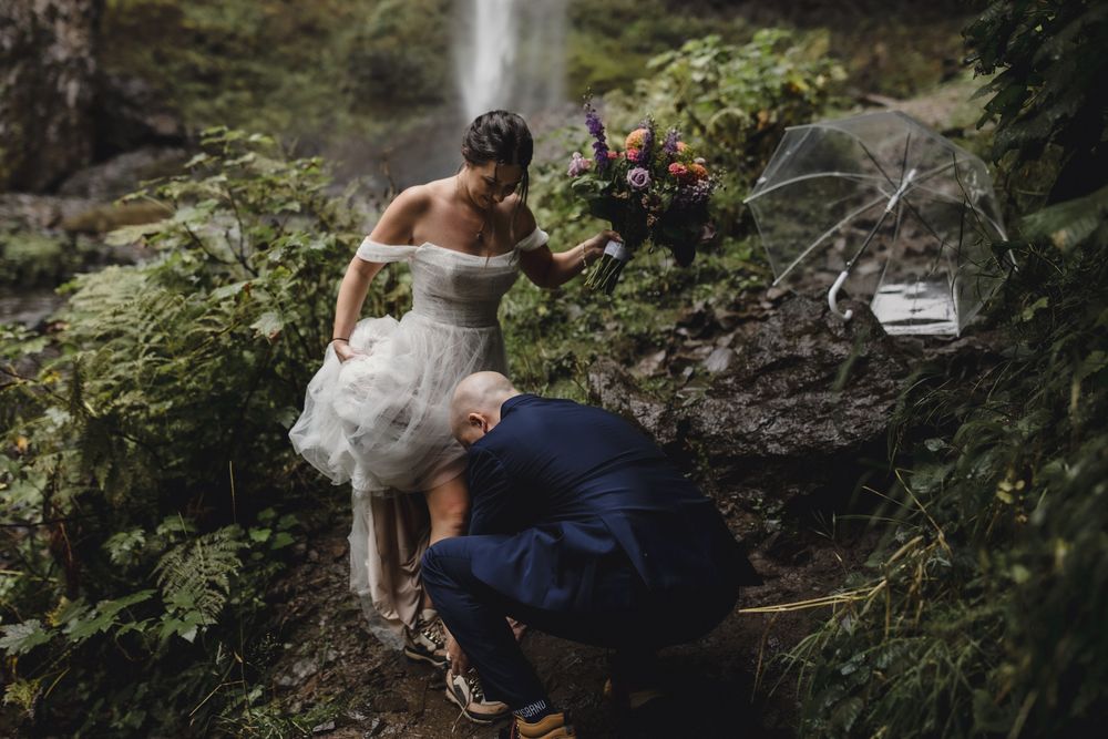Groom ties the bride's hiking boot laces before they climb up the rocks at Latourell falls in Oregon.