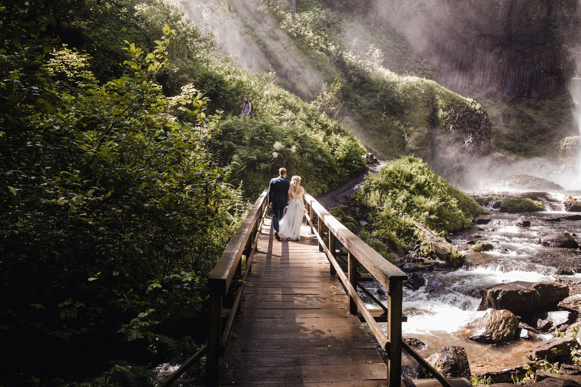 Couple walking hand-in-hand across a wooden bridge surrounded by lush green trees and a misty waterfall.