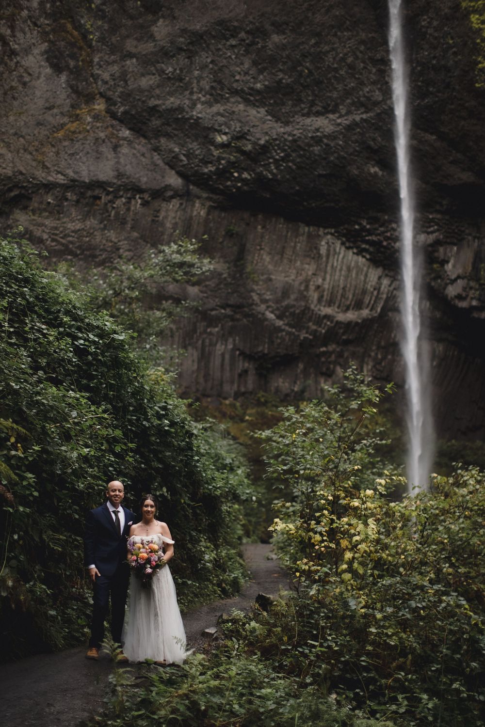 Bride and groom stand before a stunning waterfall surrounded by lush greenery, bride holding a colorful bouquet.