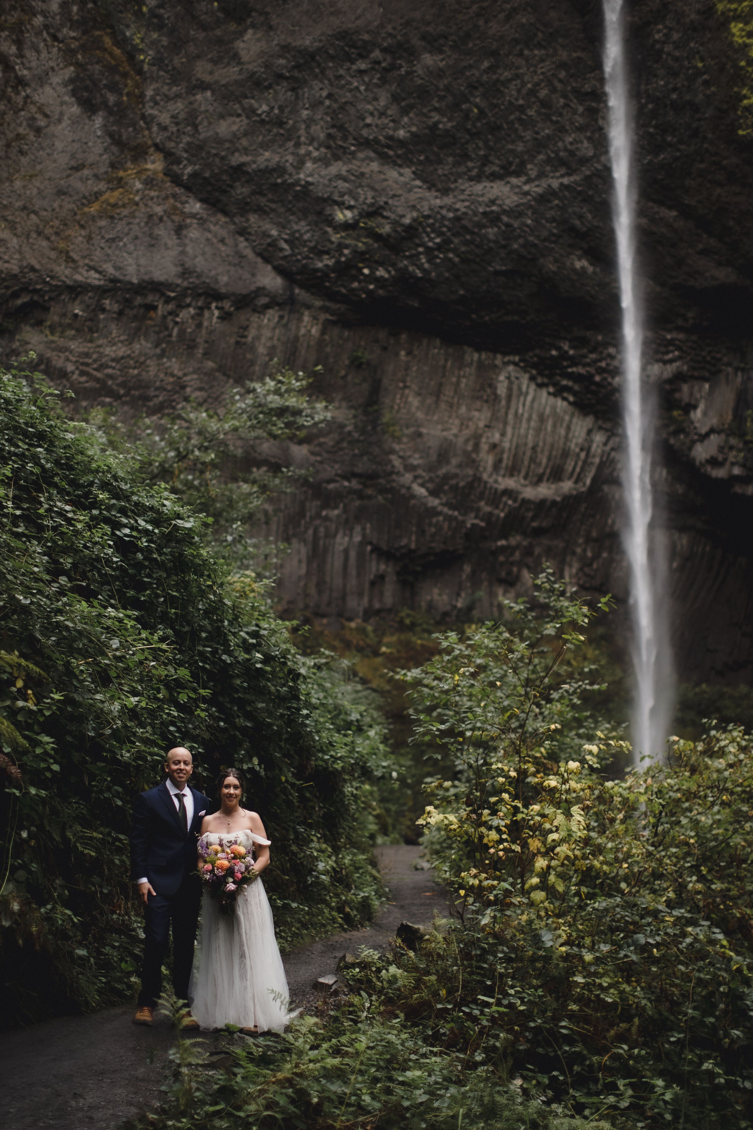 Bride and groom stand before a stunning waterfall surrounded by lush greenery, bride holding a colorful bouquet.