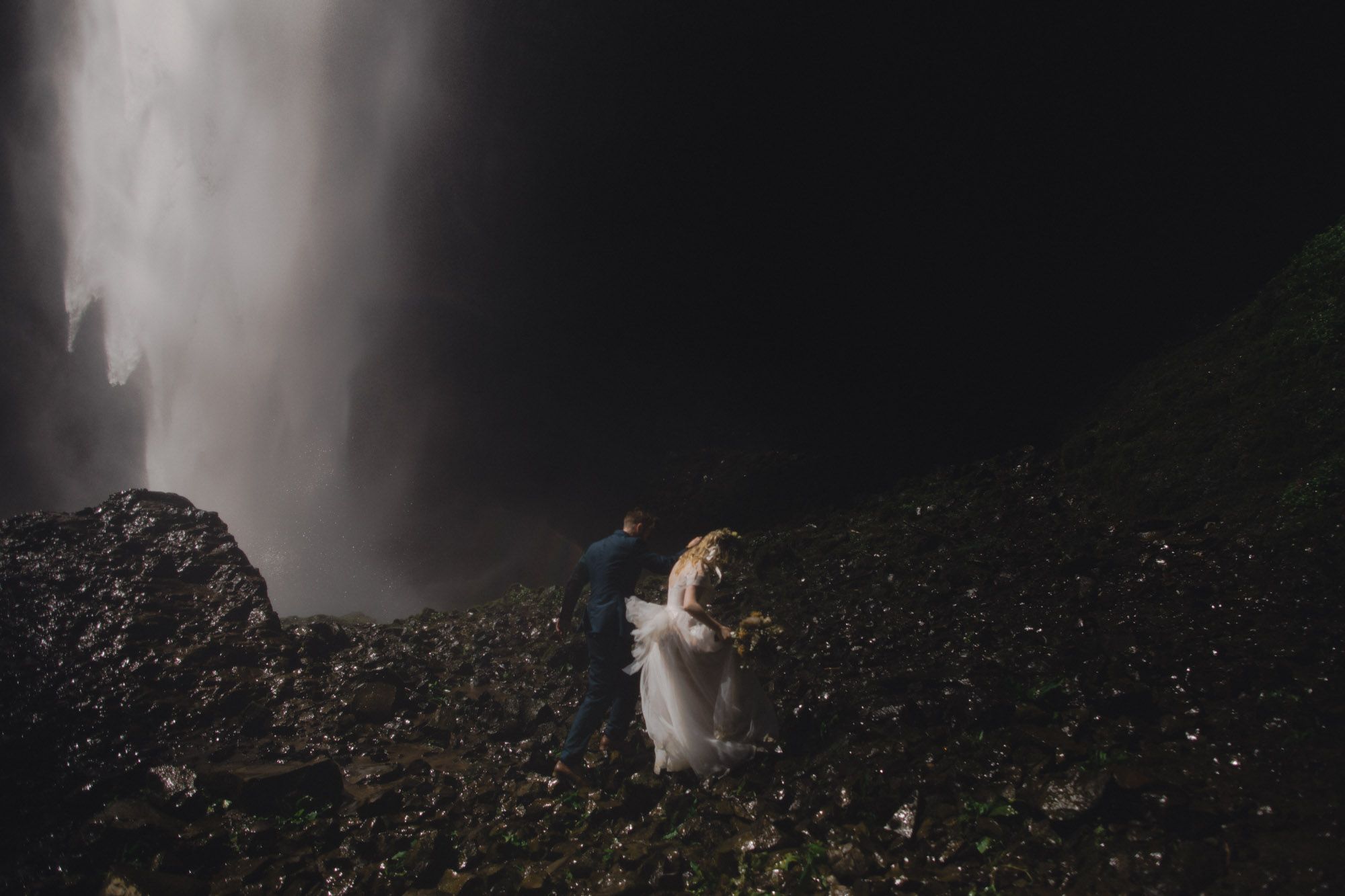 Couple walks near misty waterfall on rocky terrain during outdoor elopement in the Columbia River Gorge.