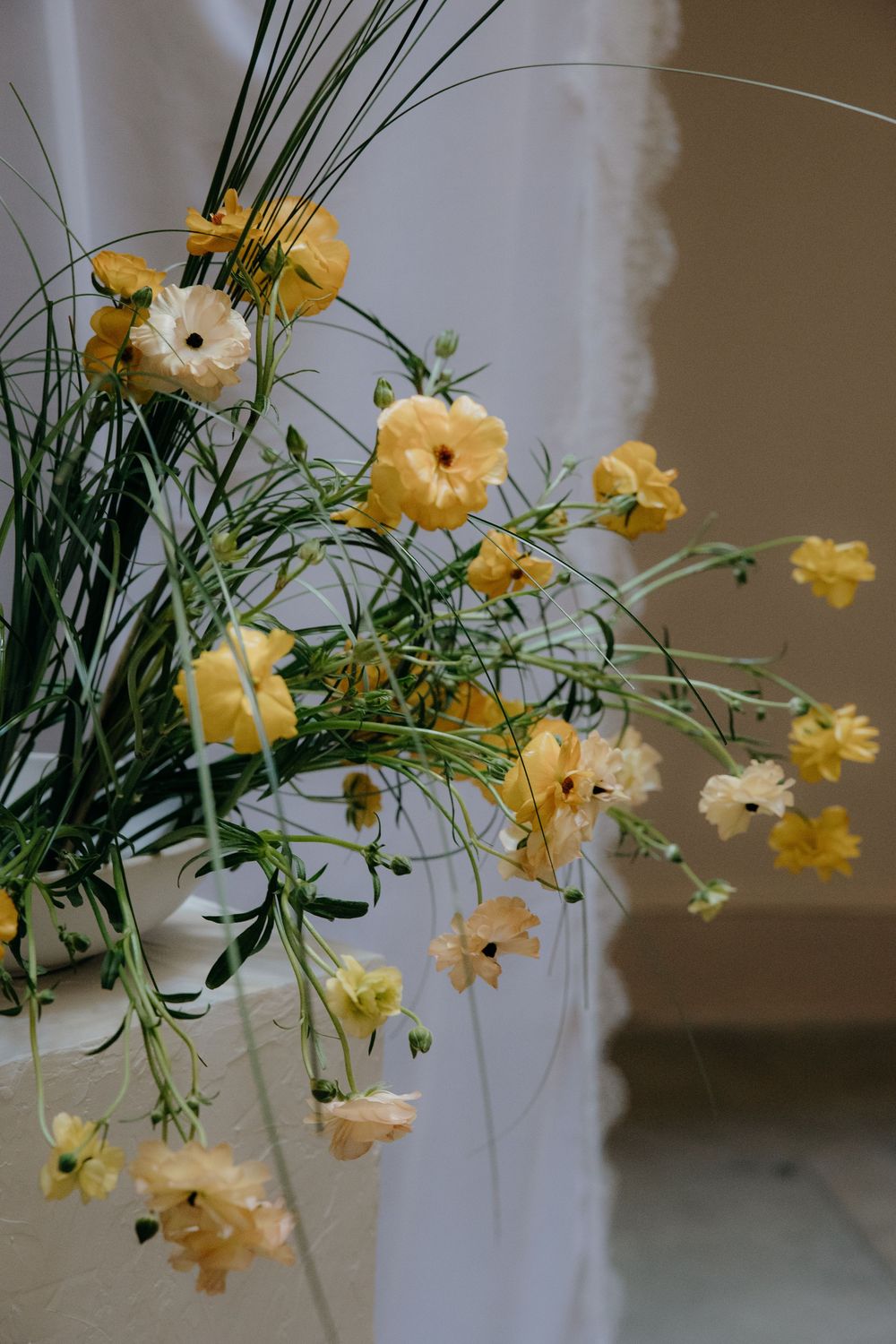 Yellow ranunculus and cosmos flowers with green grass stems arranged in a white vase against a soft white backdrop.