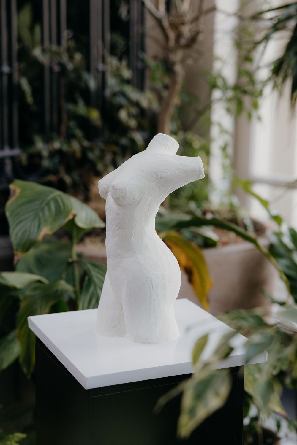 White plaster female torso sculpture displayed on a white pedestal surrounded by lush green tropical plants indoors.