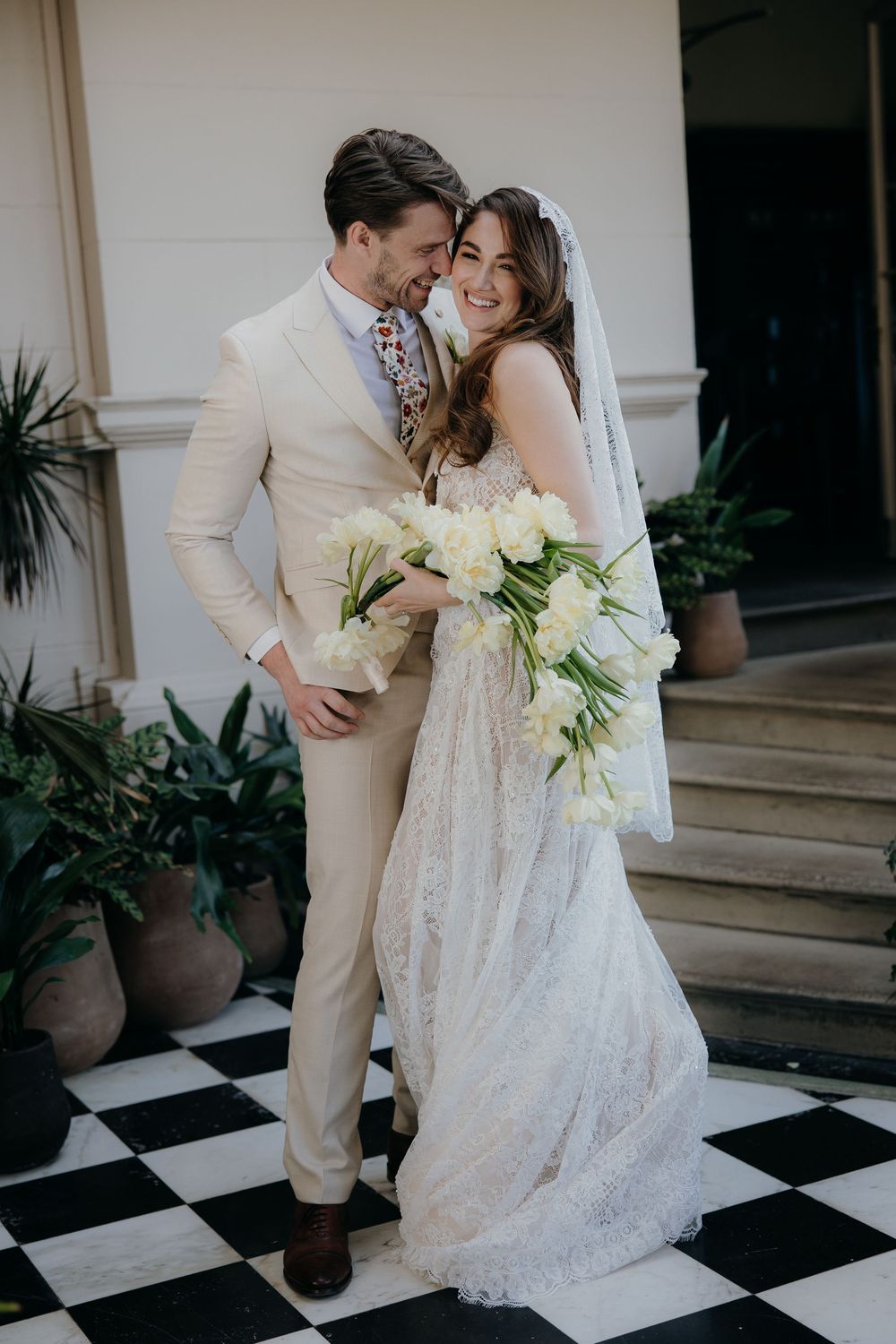 Bride in lace gown holding white peonies embraces groom in beige suit on checkered floor.