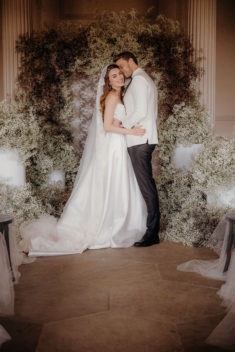Bride and groom embrace before a lush floral arch of baby's breath at their elegant wedding ceremony.