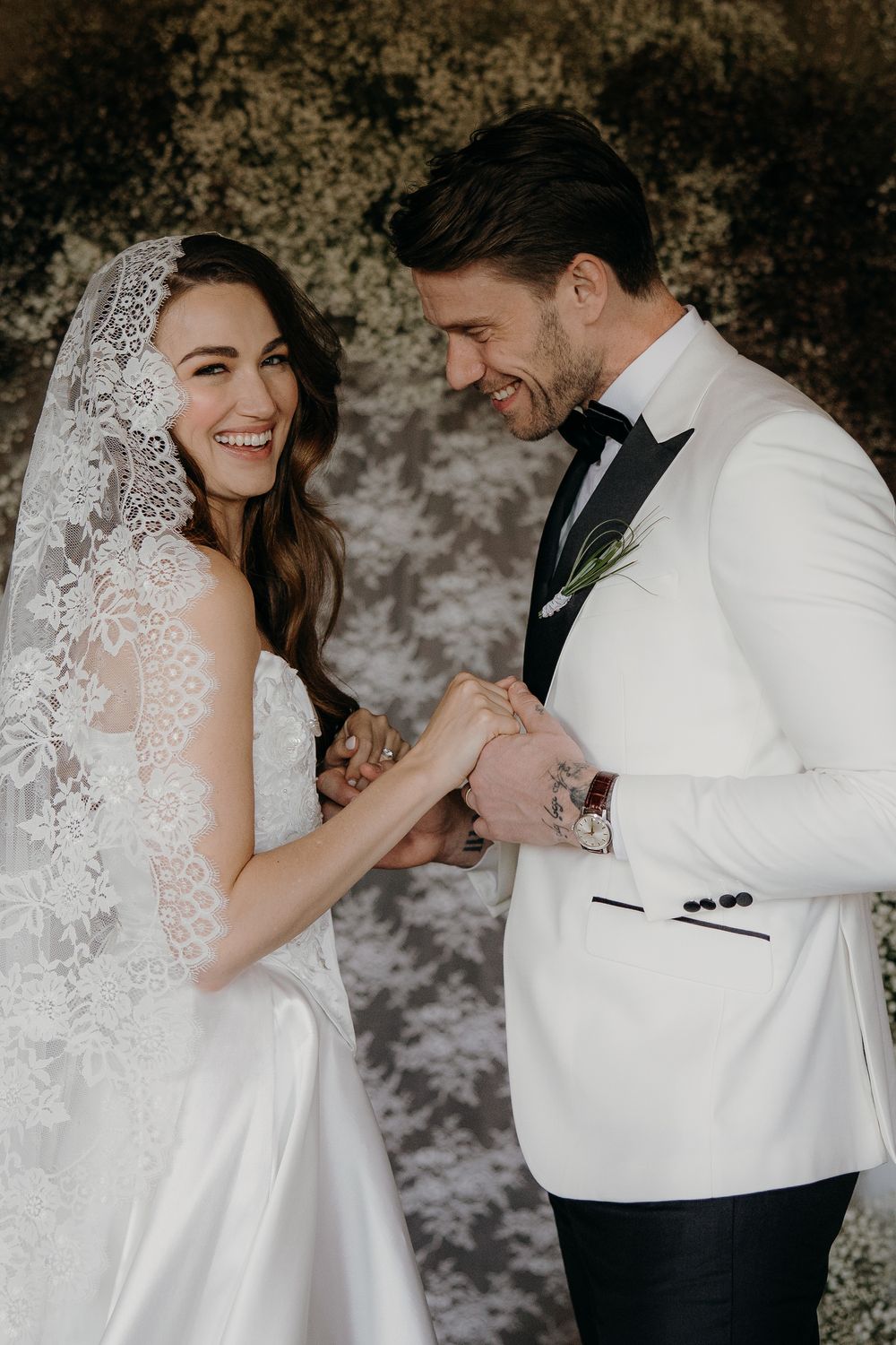 A joyful bride in lace veil and groom in white tuxedo exchange rings during an elegant wedding ceremony.