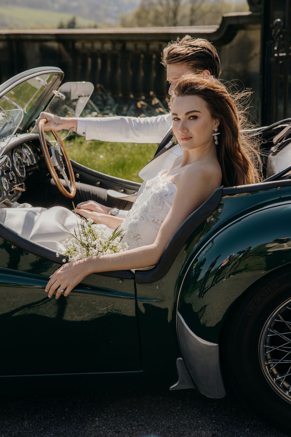 Bride in white dress sits in vintage green convertible car, holding steering wheel, with groom driving outside Denton Hall.